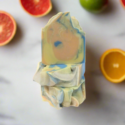 A piece of multicolored artisan soap is displayed against a simple white background. The soap features a blend of swirling colors including blue, orange, and green, resembling a marbled pattern. Its irregular, hand-crafted shape adds a unique and artistic touch, making it stand out as a decorative or functional item. The soap's design showcases a careful blending of hues, creating a visually appealing product that combines aesthetics with utility.