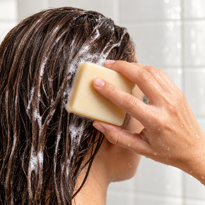 Woman holding a square pale-yellow shampoo bar against her wet brunette hair with soap lather.